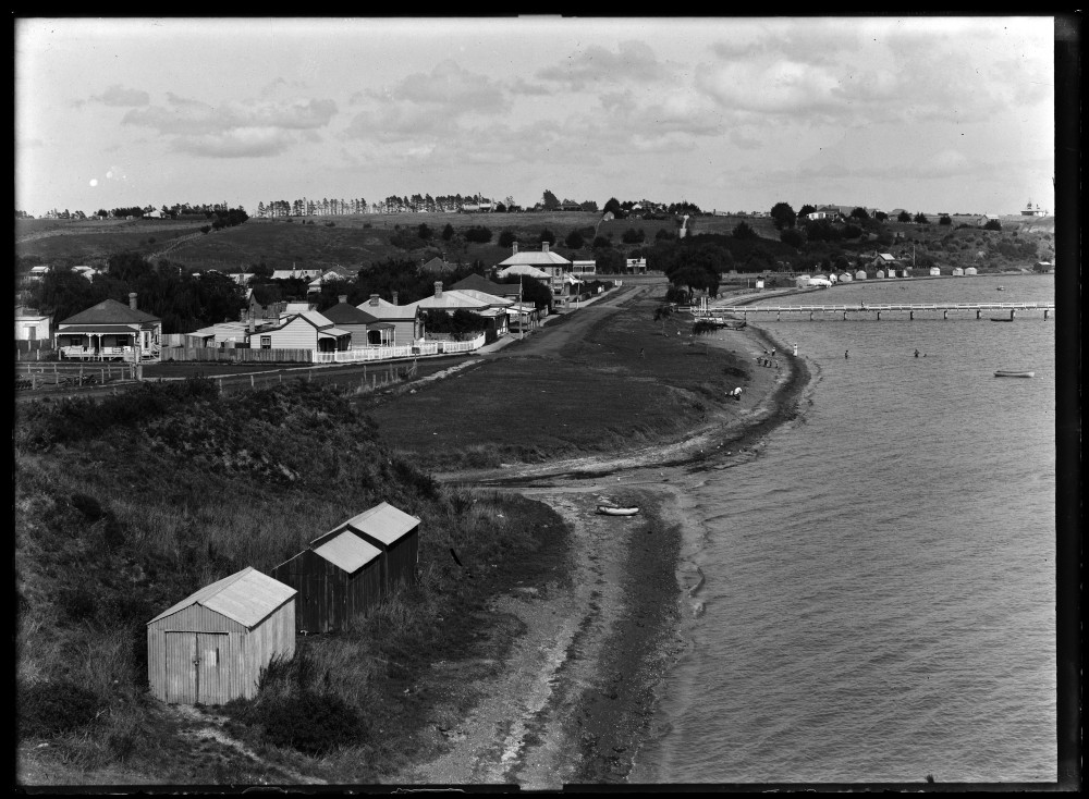 Looking west along St Heliers Bay Beach, showing St Heliers Bay Wharf, boatsheds, the intersection of Cliff Road and Tamaki Drive and St Heliers Bay Hotel